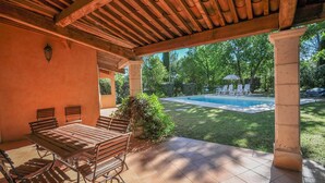 A covered patio with a wooden table and chairs, a pool in the background, and a view of the surrounding greenery. - Lovely home in Fayence (Fayence)