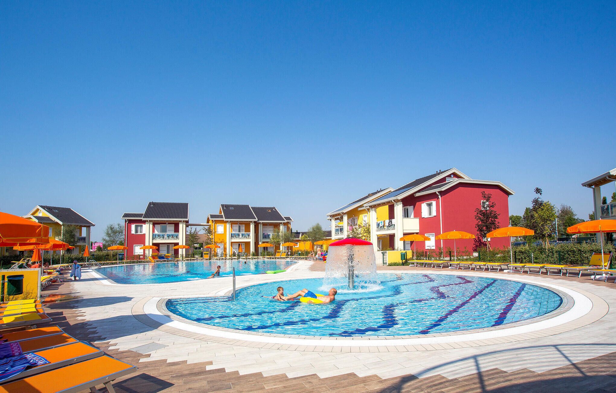 Ein Swimmingpool mit einer Fontäne, umgeben von bunten Gebäuden und orangefarbenen Sonnenschirmen.