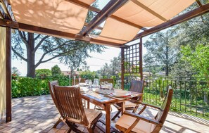 An outdoor patio with a wooden table and chairs, a pergola providing shade, and a view of greenery and trees.