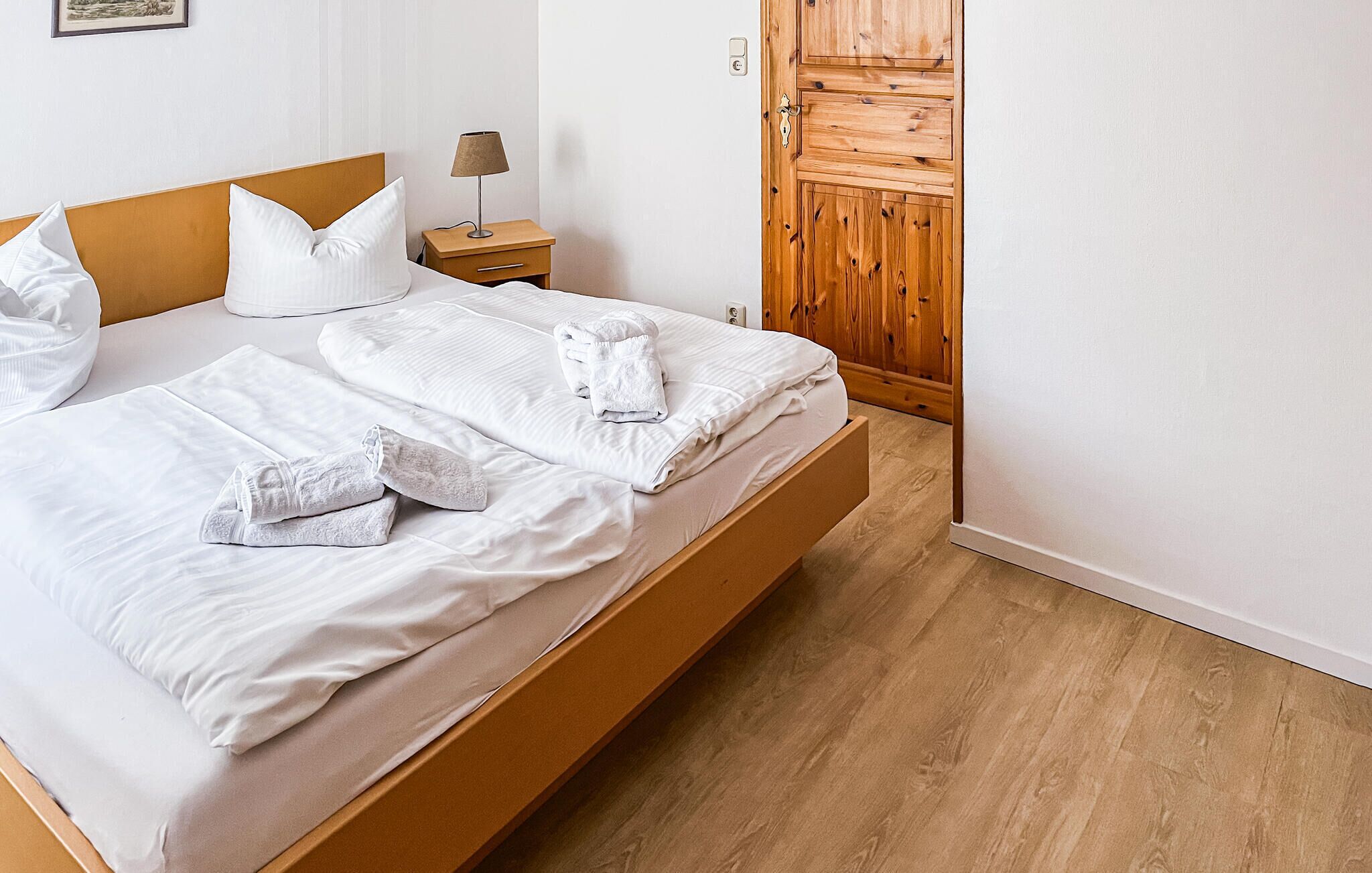 Una cama bien hecha con sábanas y toallas blancas, cabecera de madera, mesita de noche con lámpara y armario de madera.