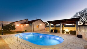 A swimming pool with stone tiles, a covered seating area, and a building with stone walls and a sloped roof. - Beach front home in Pridraga (Pridraga)