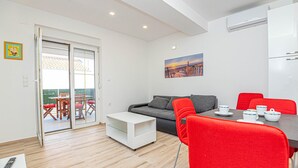 A modern living room with a sofa, a dining table with red chairs, a kitchen area, and a sliding glass door leading to an outdoor patio. - Cozy apartment in Brodarica with WiFi (Brodarica)