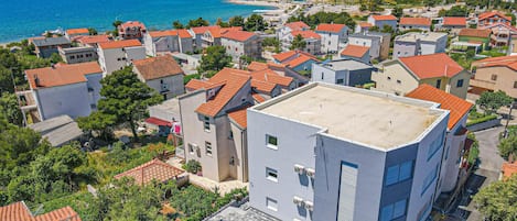 A residential area with buildings featuring red-tiled roofs, surrounded by greenery and a coastal view.