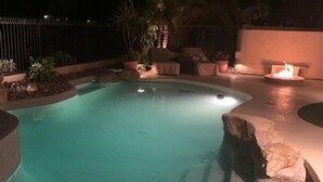 Una piscina iluminada con luz nocturna, de borde curvo, rodeada por un área ajardinada con rocas y plantas.