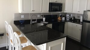 Una cocina moderna con armarios blancos, encimera negra, electrodomésticos de acero inoxidable y una zona de comedor con sillas blancas.