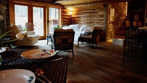 Ein rustikales Blockhaus-Interieur mit Holzboden, einer Couch, zwei Sesseln, einem Esstisch mit Tellern und einem Löffel sowie einer Lampe.