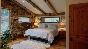 Ein Schlafzimmer mit Steinwand, Holzbalken, einem großen Bett, zwei Nachttischlampen und Blick ins Freie.