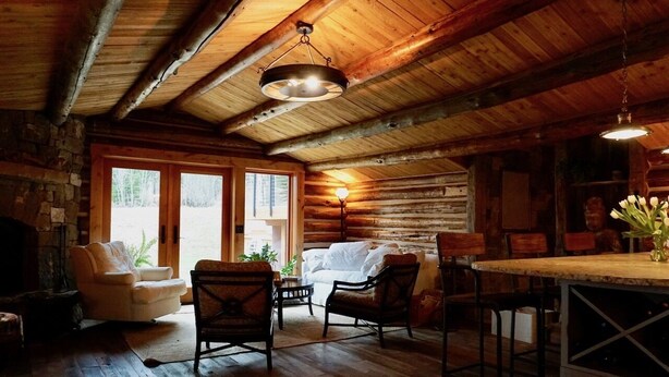 Ein rustikales Blockhaus-Interieur mit Holzbalkendecke, Kamin, einer Couch und einem Essbereich mit Stühlen.