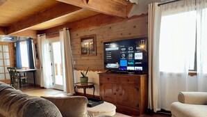 Ein gemütliches Wohnzimmer mit Holzboden, einem Flachbildfernseher, der eine Streaming-Oberfläche anzeigt, einem Sofa, einem kleinen Tisch und einem Fenster mit Vorhängen.