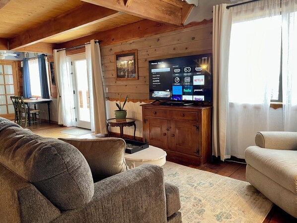 Una sala de estar acogedora con techo de madera, un televisor de pantalla plana mostrando la interfaz de un servicio de streaming, un sofá, una mesita y una ventana con cortinas.