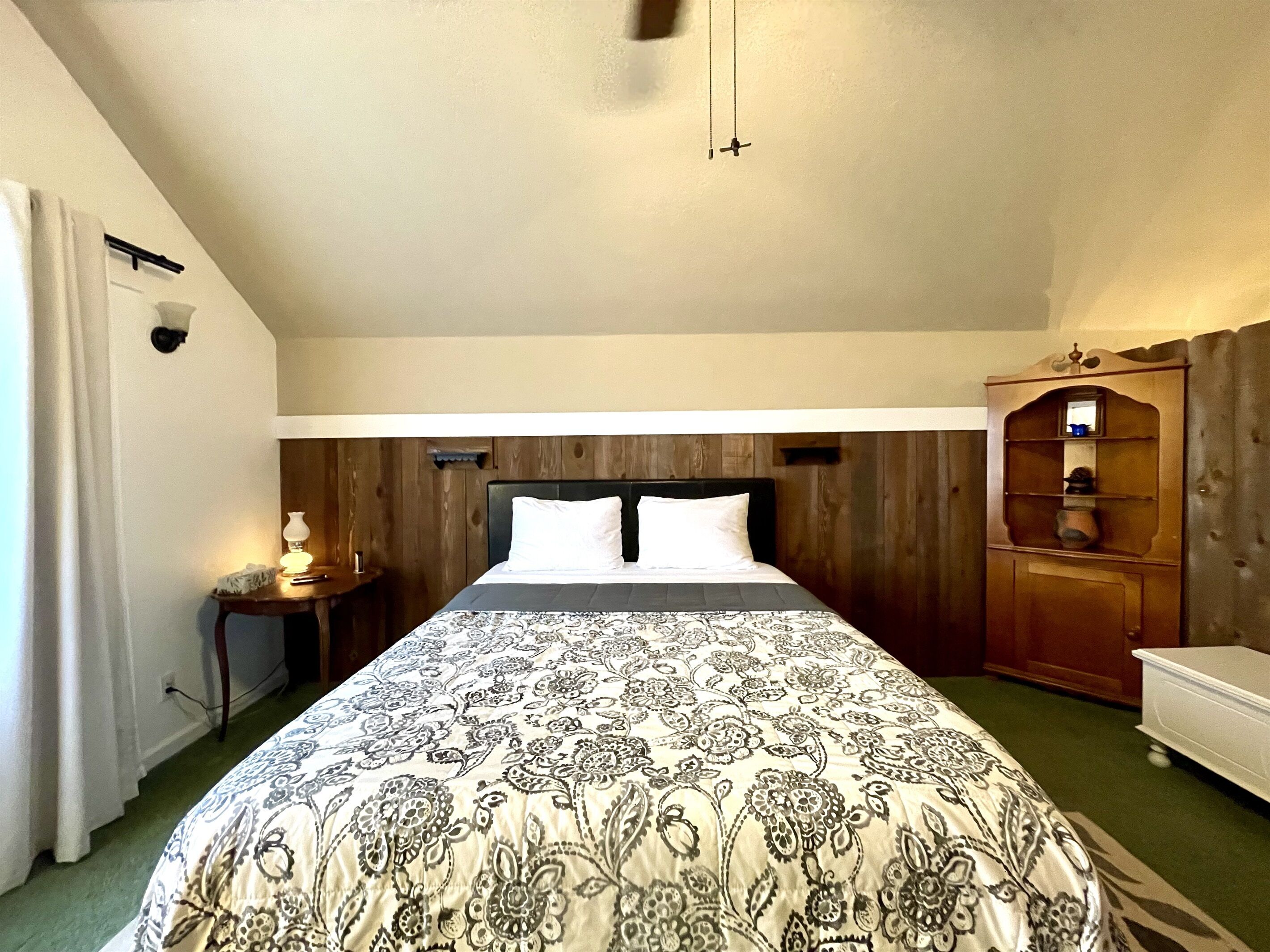 A bedroom with a large bed, wooden paneling on the walls, a bedside table with a lamp, and a wooden cabinet.