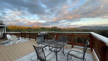A deck with outdoor furniture, including chairs and a table, overlooking a scenic landscape with trees and mountains.