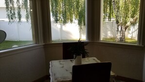 A room with a table and chair, a window with a view of a white fence and trees, and a hanging plant.