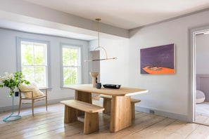 A dining area with a wooden table and benches, a hanging light fixture, and a painting on the wall.