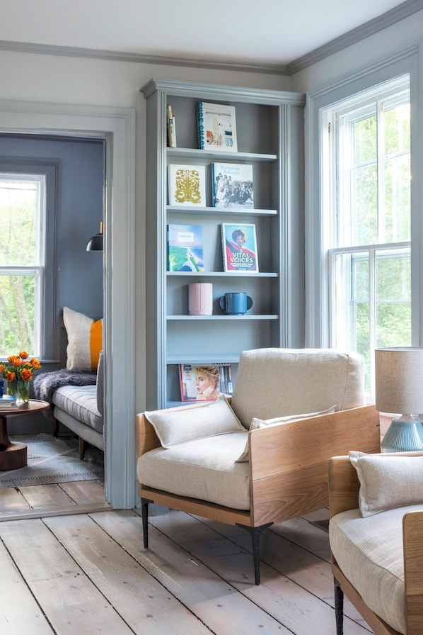 A cozy room with a wooden floor, a bookshelf, a comfortable chair, and a window with a view of trees.