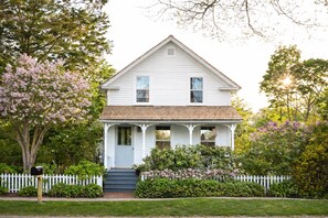 A two-story white house with a gabled roof, a front porch with columns, and a white picket fence.