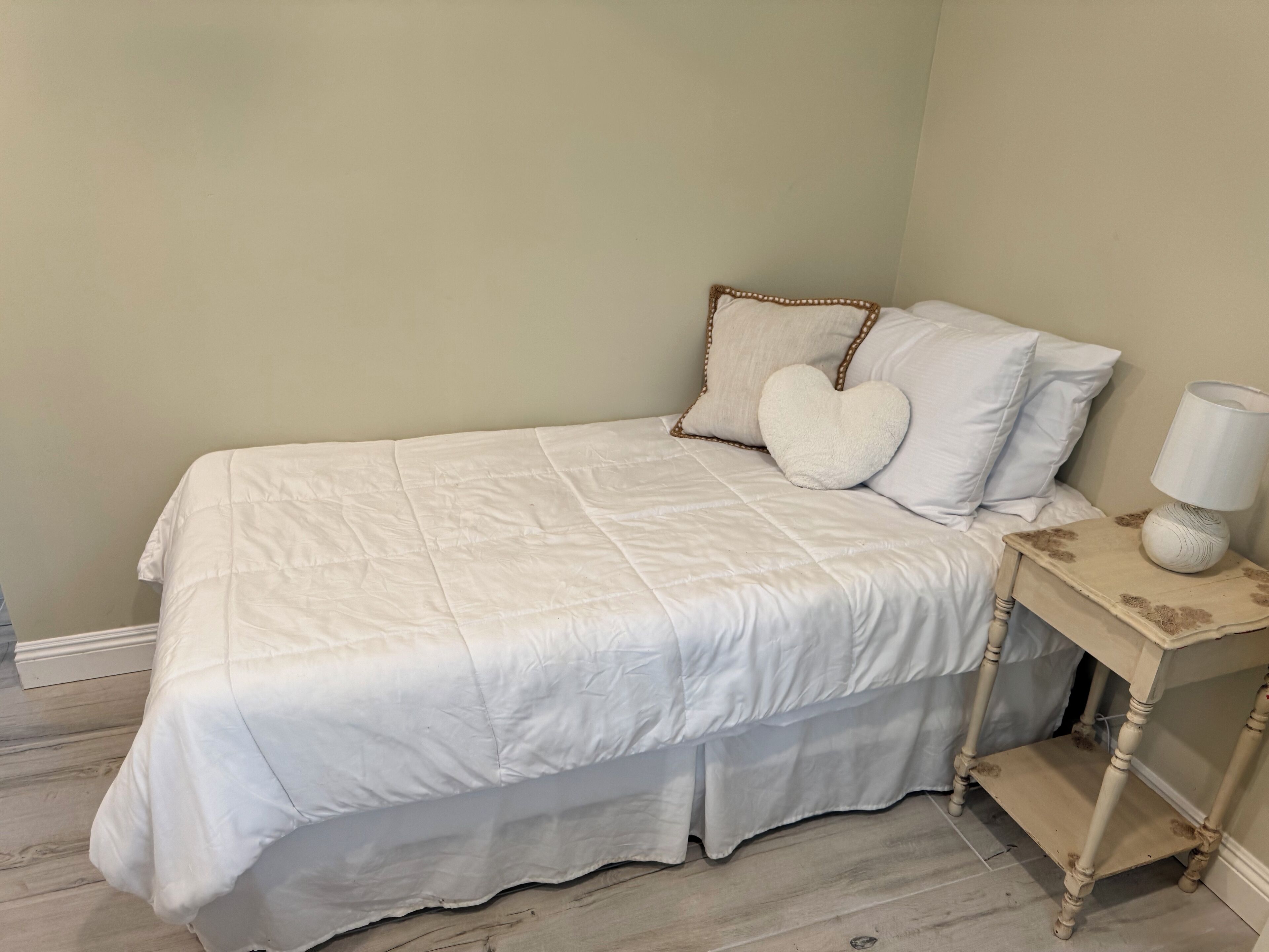 Un lit bien fait, recouvert d’une couette blanche, avec deux oreillers en forme de cœur et une table de chevet sur laquelle se trouve une lampe.