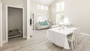 A spacious room with a dining table, chairs, a sofa with colorful pillows, and a view of the outdoors through large windows. - Adorable 4 Bedroom Guest House wPool + Jacuzzi (Poway)