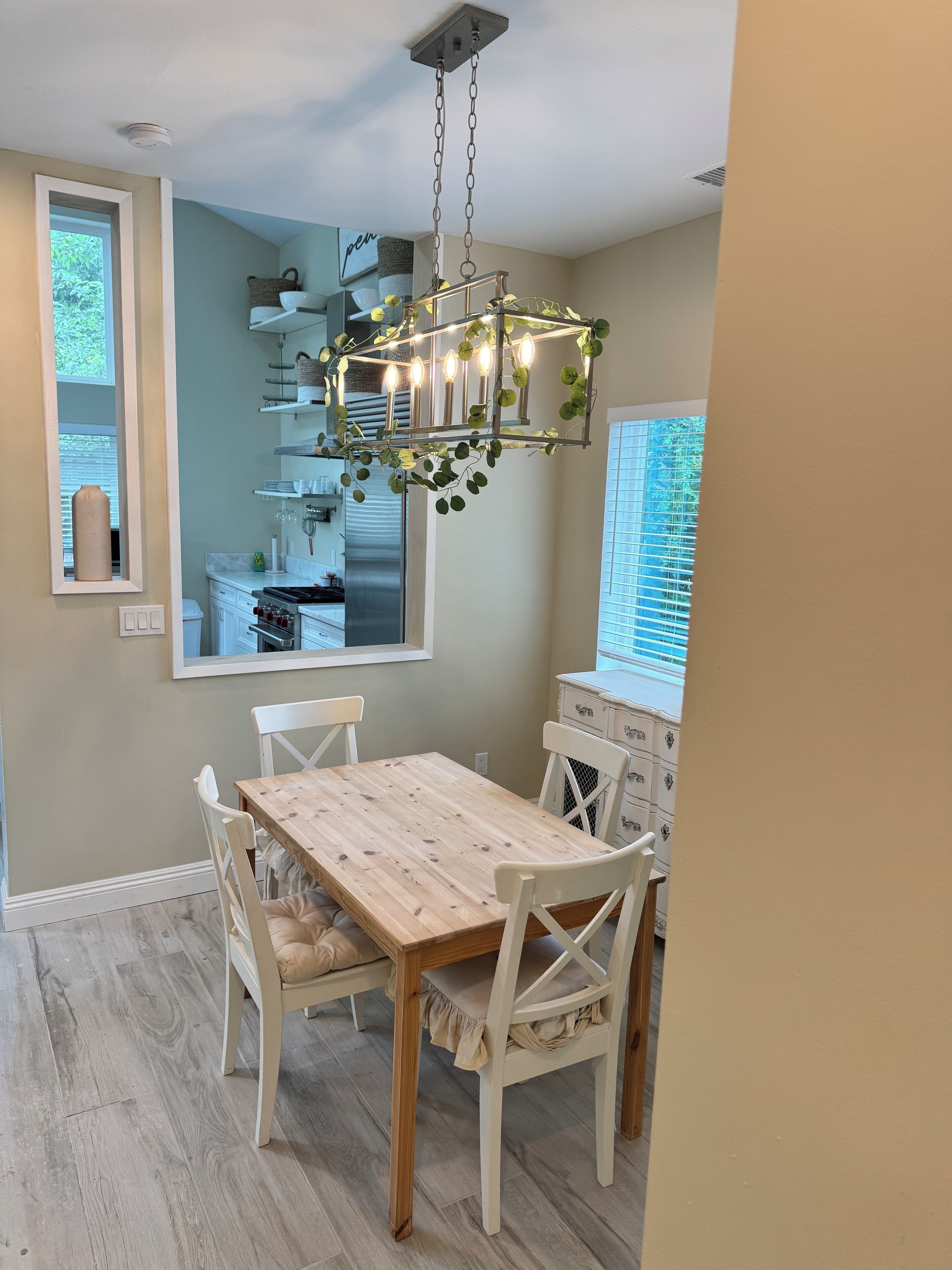 A dining area with a wooden table and white chairs, a hanging light fixture with greenery, and a view of the kitchen through a window.