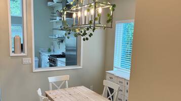 A dining area with a wooden table and white chairs, a hanging light fixture with greenery, and a view of the kitchen through a window.