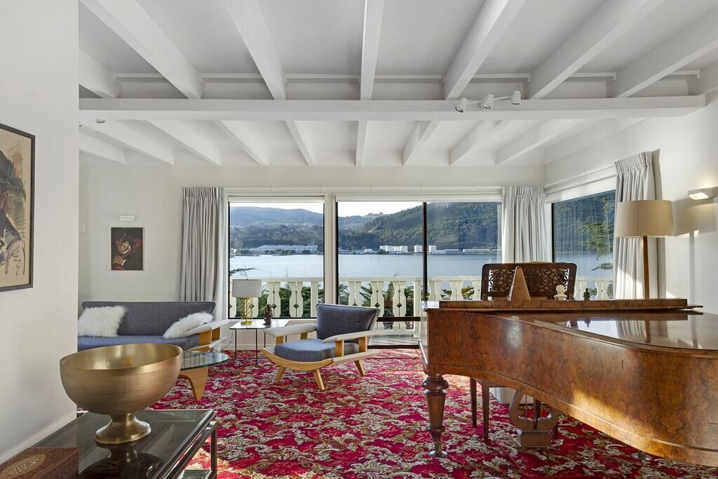 A spacious room with a grand piano, a large window offering a scenic view of a lake and mountains, and a red patterned carpet.