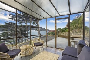 A modern room with large glass windows, wicker furniture, and a view of a coastal landscape. - Harbour Views (Dunedin)