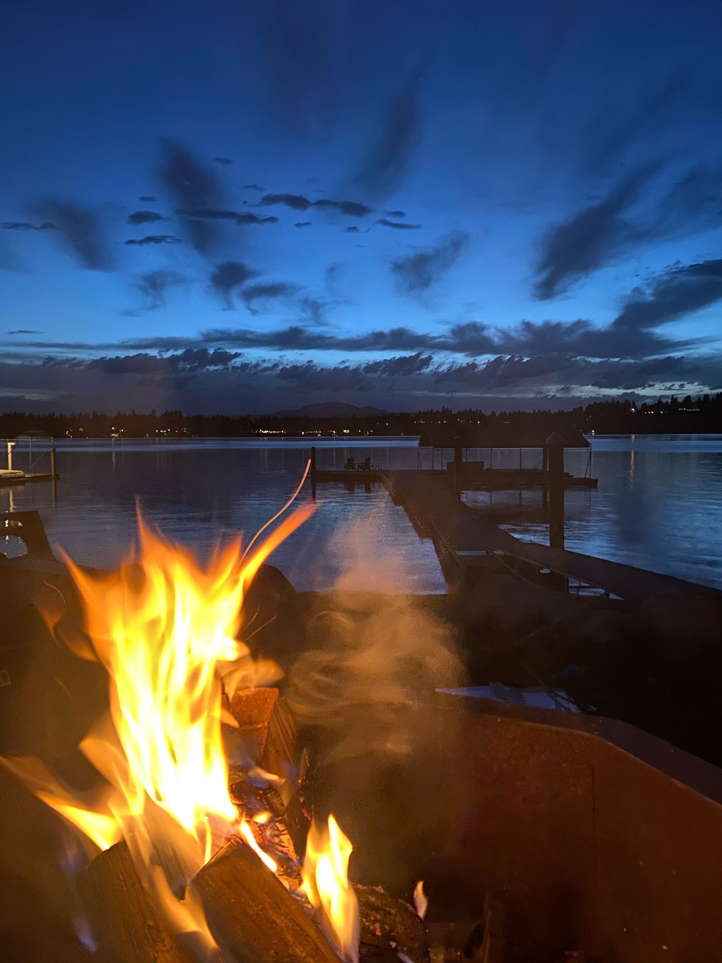 A fire burning on a dock with a calm lake and mountains in the background.