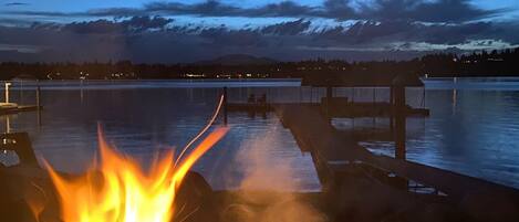 A fire burning on a dock with a calm lake and mountains in the background.