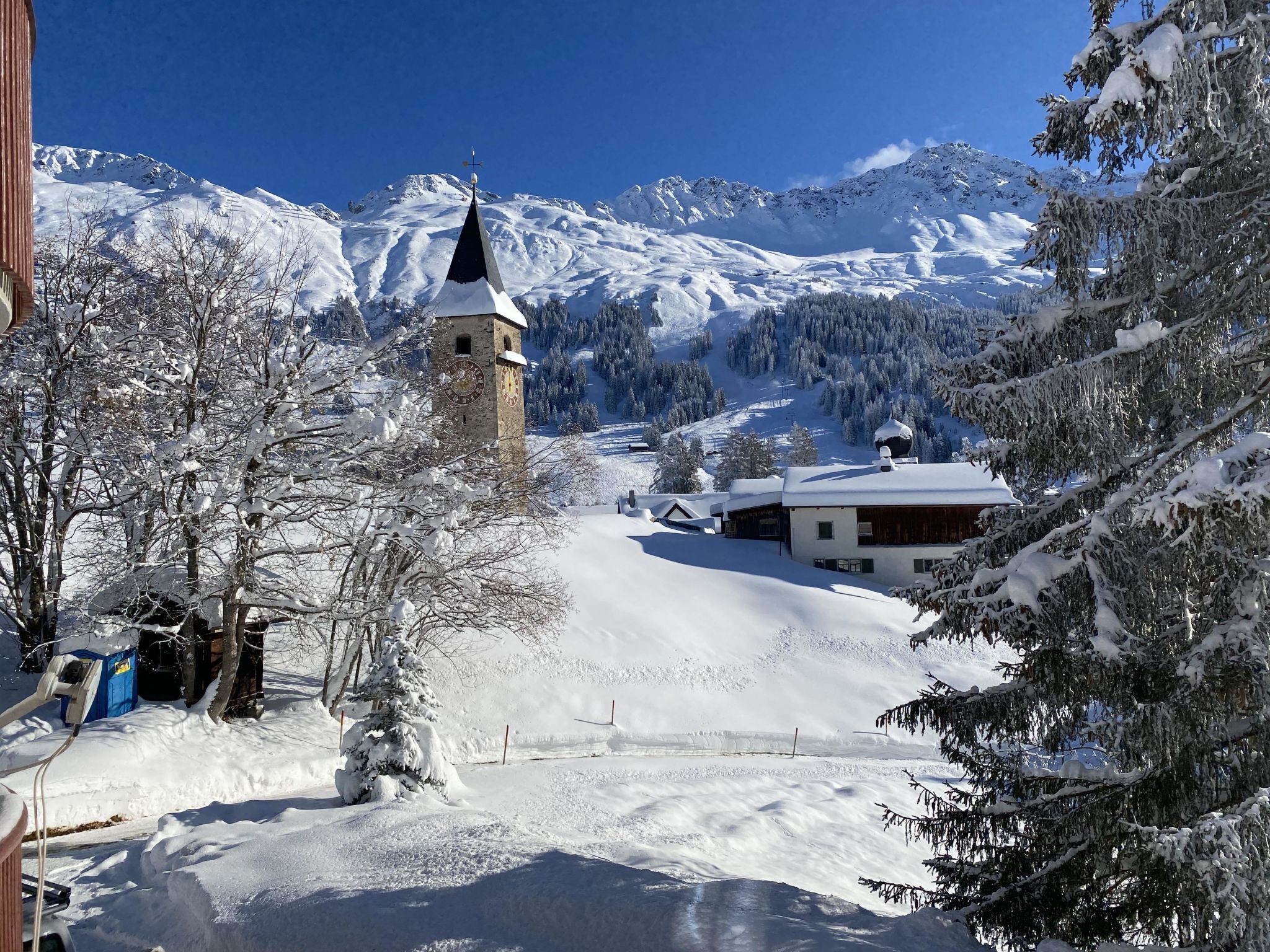 A snow-covered village with a church tower, a wooden building, and a ski slope in the background.