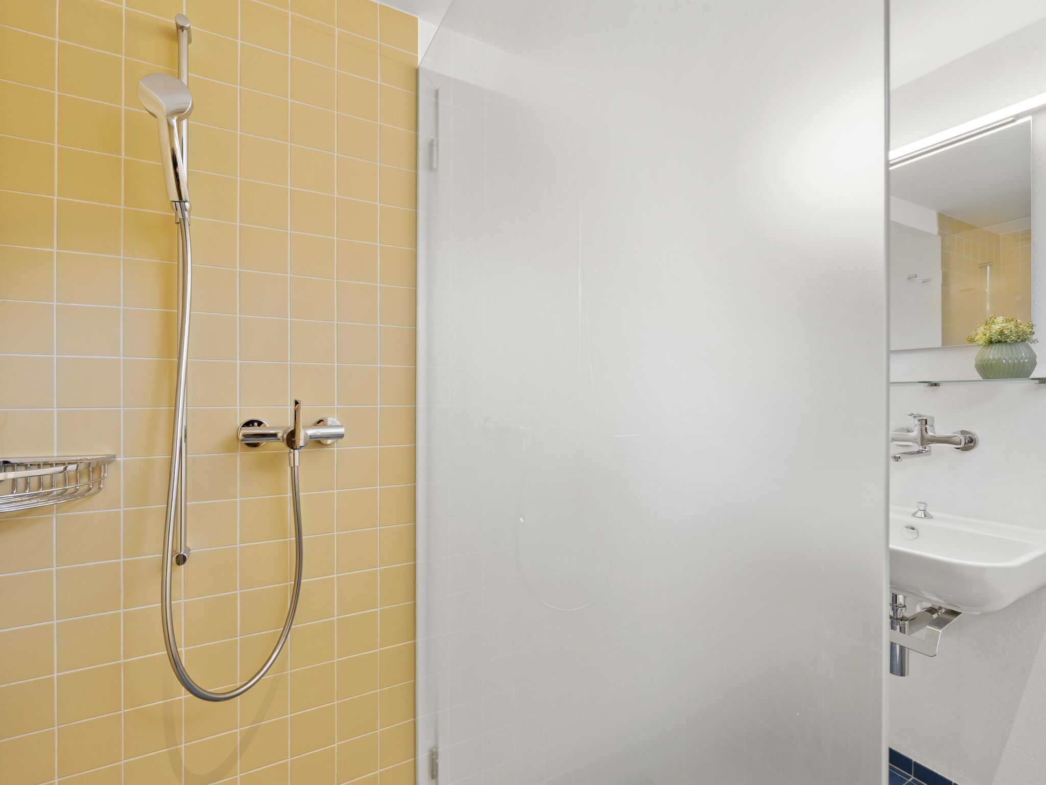 Un baño con paredes de azulejos amarillos, una ducha con cabezal de mano, cerramiento de ducha de vidrio y un lavabo blanco con un espejo encima.