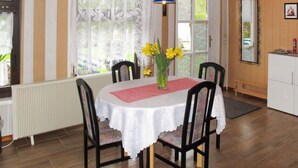 Un comedor con una mesa redonda, sillas, un jarrón con flores, una ventana con cortinas, un radiador y un armario con un espejo.