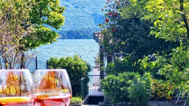 Un jardín con vistas a un lago y montañas, dos copas de vino sobre una mesa y un sendero que lleva a un edificio.