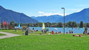 Un parque junto al lago con zonas verdes, un velero y montañas al fondo.