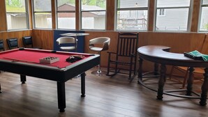 Une table de billard entourée de chaises, une petite table avec une chaise, et de grandes fenêtres donnant sur l’extérieur.