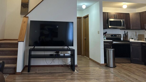 Un salon moderne doté d’un téléviseur à écran plat, d’un escalier en bois et d’une cuisine avec des meubles sombres et des appareils électroménagers en acier inoxydable.