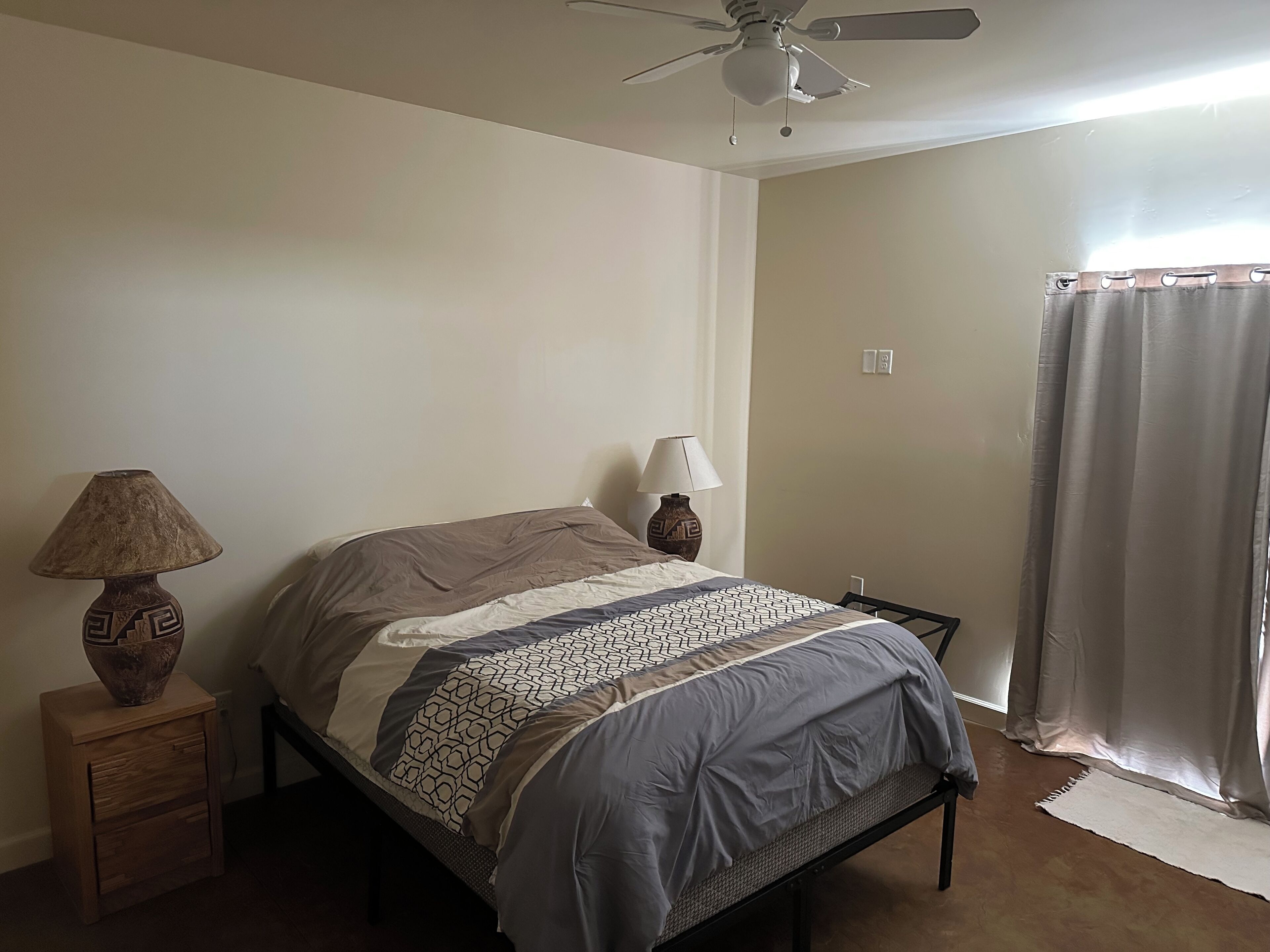 Une chambre à coucher avec un lit, deux lampes de chevet, un ventilateur de plafond et une fenêtre avec des rideaux.