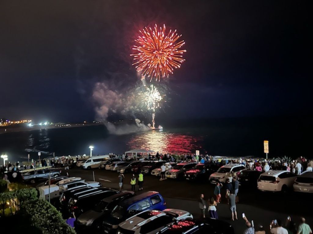 Un événement nocturne en plein air, avec un feu d’artifice tiré au-dessus de l’eau, une foule et des voitures garées.