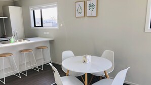 Une cuisine moderne avec un plan de travail blanc, des tabourets en bois et une table ronde entourée de chaises blanches. Des tableaux encadrés ornent le mur, et une fenêtre se trouve au-dessus de l’évier.