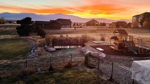 A fenced outdoor area with a playground, including a slide and a swing set, surrounded by a grassy field and a mountain range in the background. - Massive Spacious House with Views, Adventure and Location (Colorado Springs)