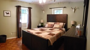 A bedroom with a large bed, two bedside tables with lamps, a wooden headboard, a window with curtains, a wall-mounted deer skull, and a framed picture on the wall.