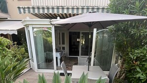 A covered patio area with a table and chairs, surrounded by lush greenery and a building with a balcony. - Mistral Gagnant (Fréjus)