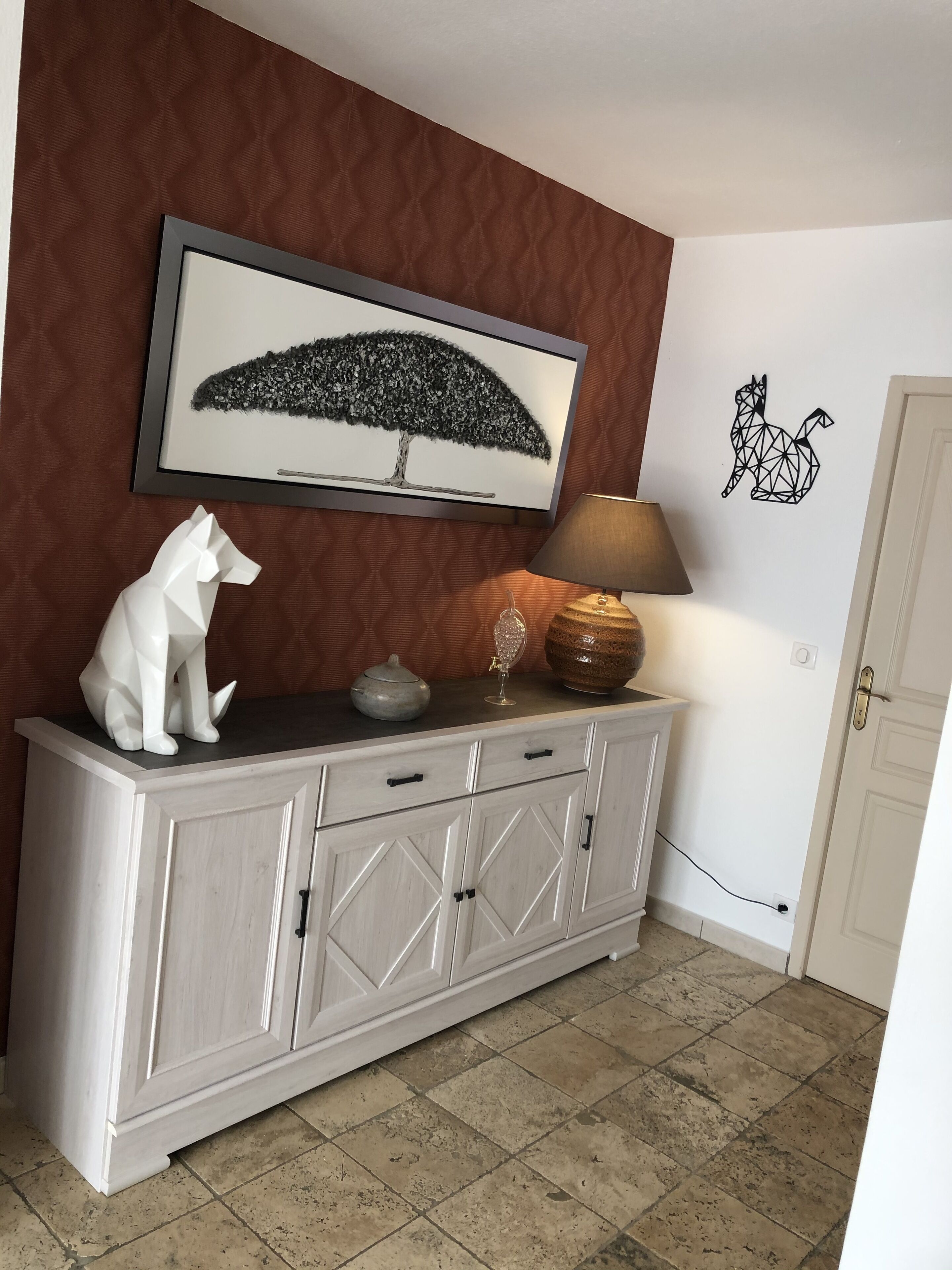 Un meuble blanc avec un plateau orné d’un motif géométrique, un tableau encadré représentant un arbre, une lampe et un autocollant mural représentant un chat.
