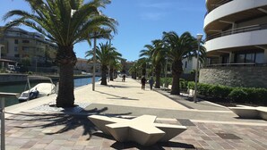 A waterfront promenade with palm trees, a boat dock, and a modern building with balconies. - Mistral Gagnant (Fréjus)
