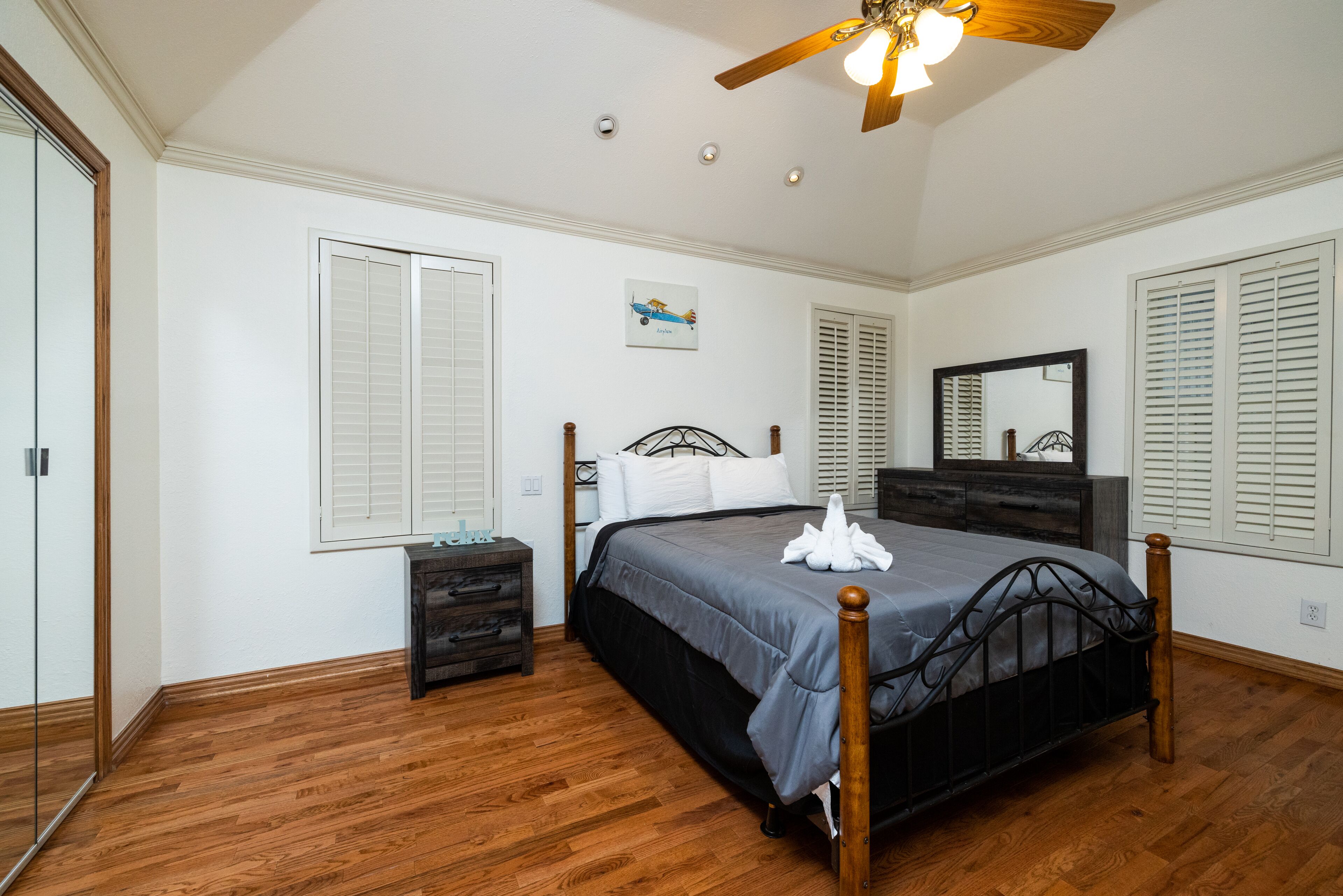 A bedroom with a wooden floor, a bed with a gray bedspread, a nightstand, a dresser, and a ceiling fan.