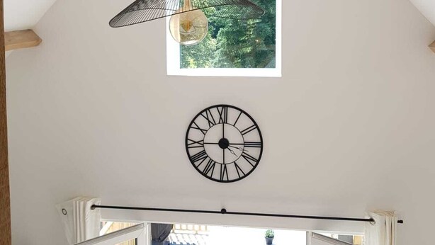 A room with a sloped ceiling, a hanging pendant light, a wall clock, and a window with a view of greenery.