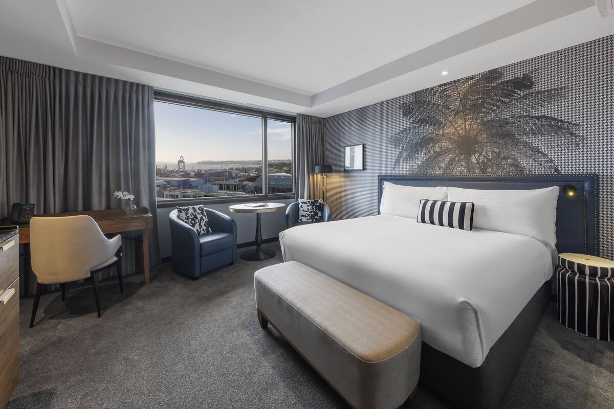 A hotel room with a large bed, a desk, a chair, a small table, and a view of the city through a large window.