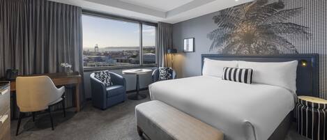 Habitación de hotel con una cama grande, un escritorio, una silla, una mesita y vistas a la ciudad a través de un ventanal.