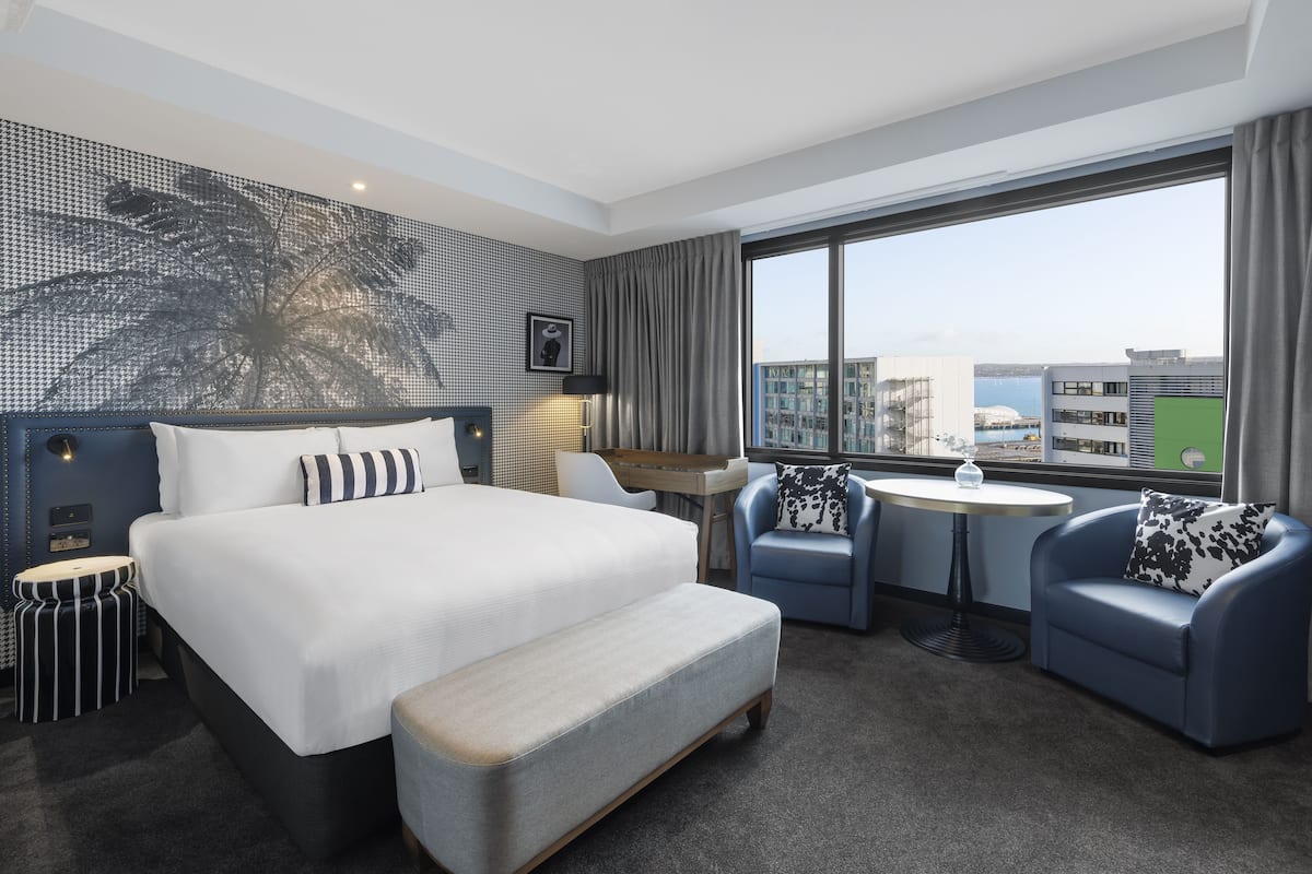 A modern hotel room with a large bed, two armchairs, a small table, and a view of the city.