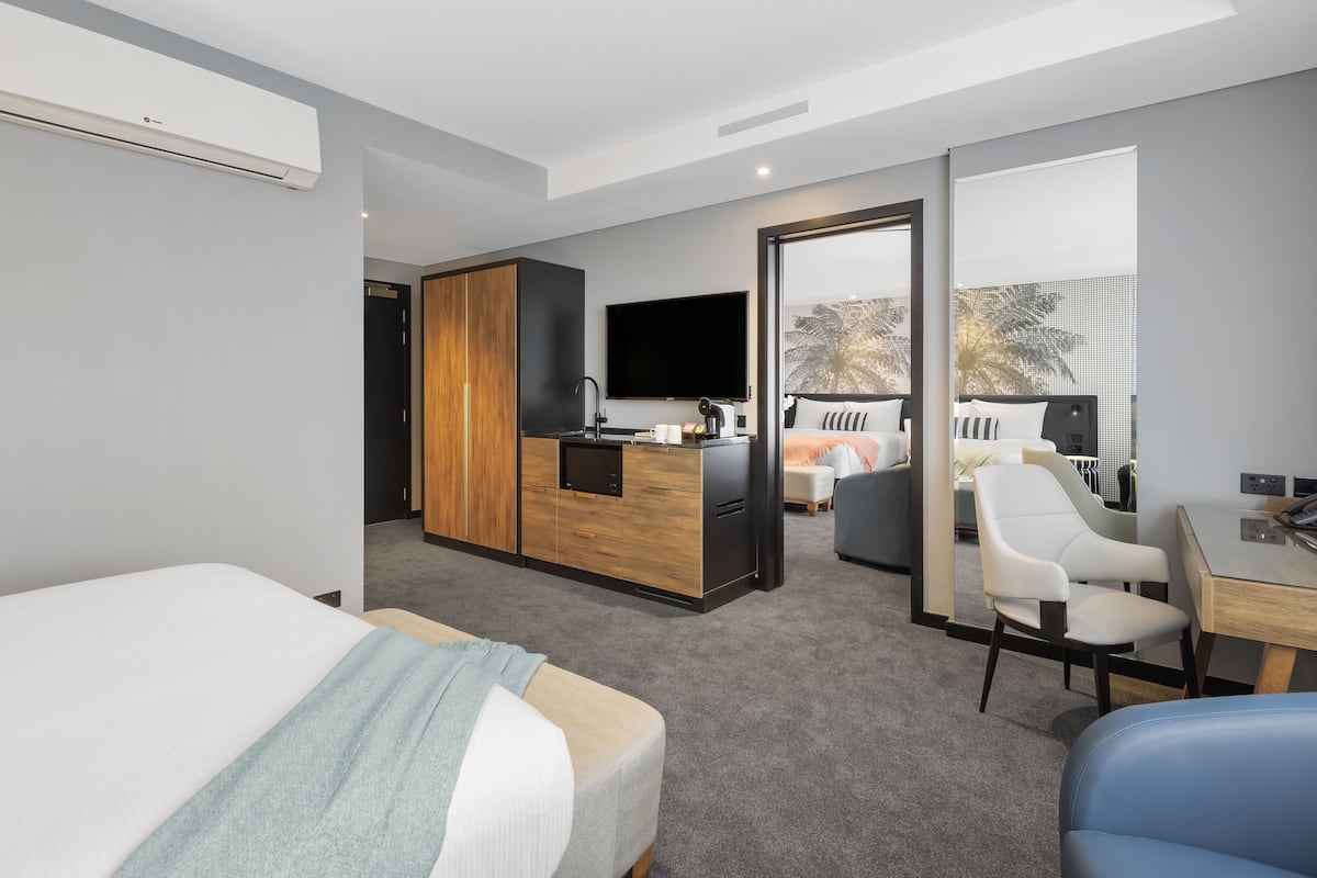 A modern hotel room with a bed, a desk, a chair, a kitchenette, and a TV.