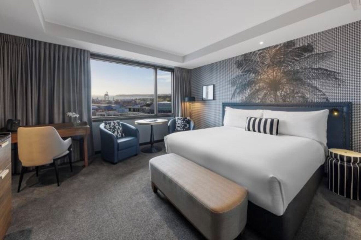 Habitación de hotel moderna con una cama grande, un escritorio, dos sillas, una mesita pequeña y vistas de la ciudad a través de amplias ventanas.